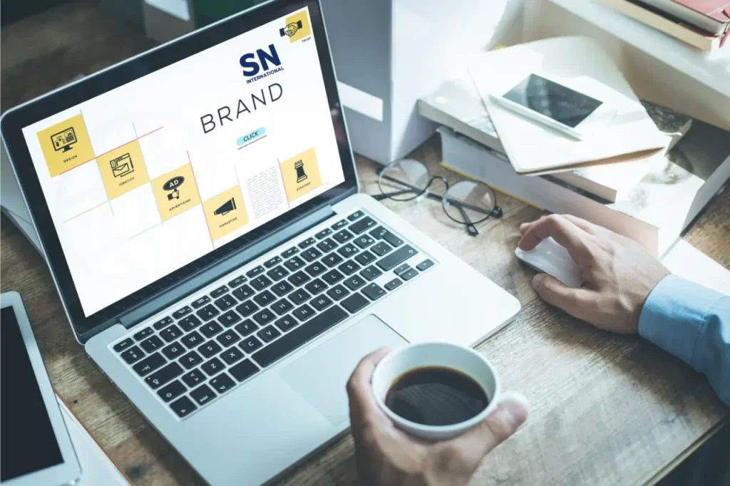 A person working on a laptop displaying a custom branding design, holding a cup of coffee. The SN International logo is visible on the screen.