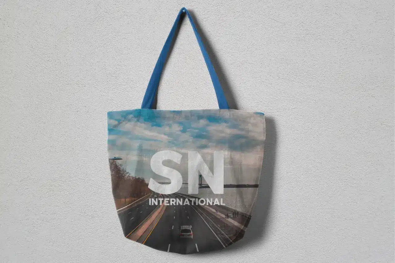 A stylish tote bag with a scenic road design featuring a clear sky and a distant bridge. The bag prominently displays the logo 'SN INTERNATIONAL' in bold white letters. The handles are a solid blue color, adding a pop of contrast to the muted tones of the bag's design.