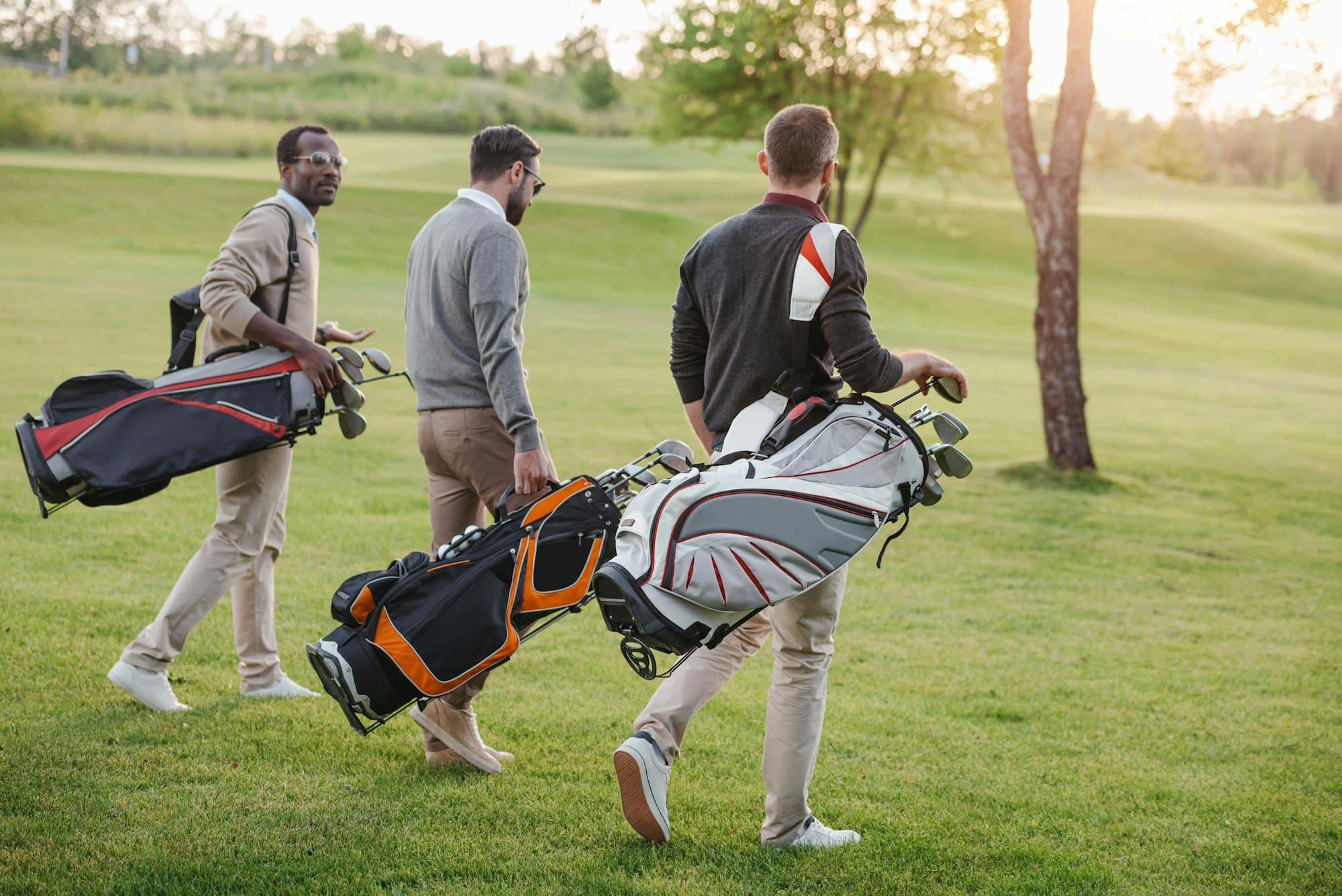 multiethnic golf players with golf clubs in bags walking on golf course