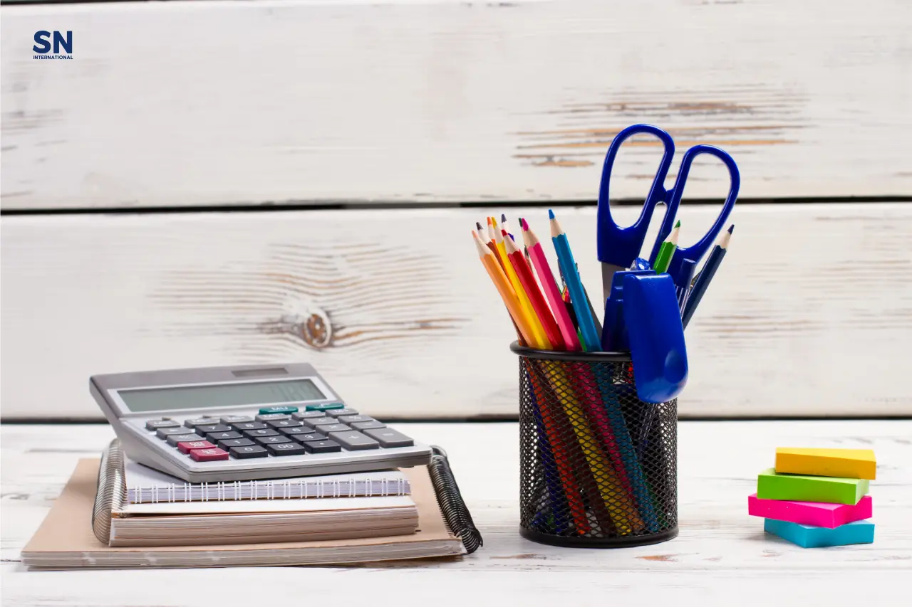 A neatly arranged desk setup with a calculator on top of stacked notebooks, a pen holder containing colorful pencils, blue scissors, and other office supplies. The SN INTERNATIONAL logo is visible in the top left corner.